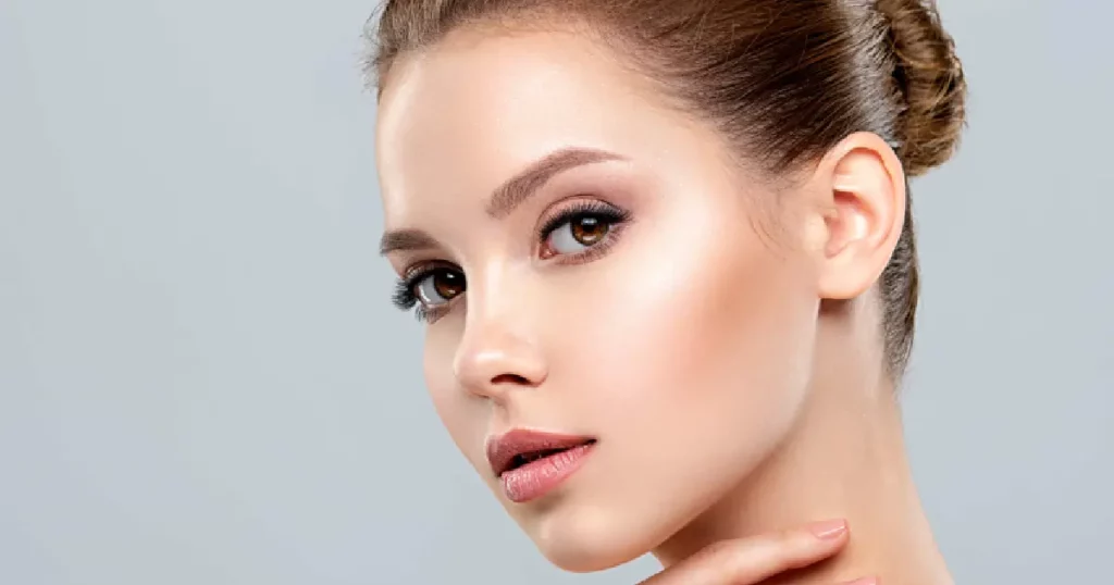 Young woman with flawless, glowing skin, styled hair in a neat bun, and a hand gently touching her neck, against a light gray background, representing Skinvive in Northborough, MA.